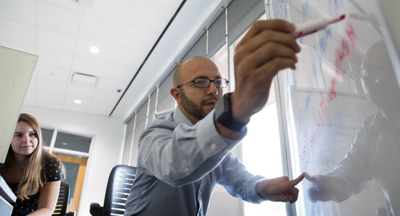 Professor writes on white board while student listens.