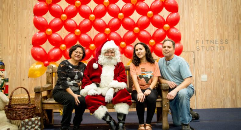 Santa Claus, a student and their family sit for a photo in front of a red balloon backdrop.