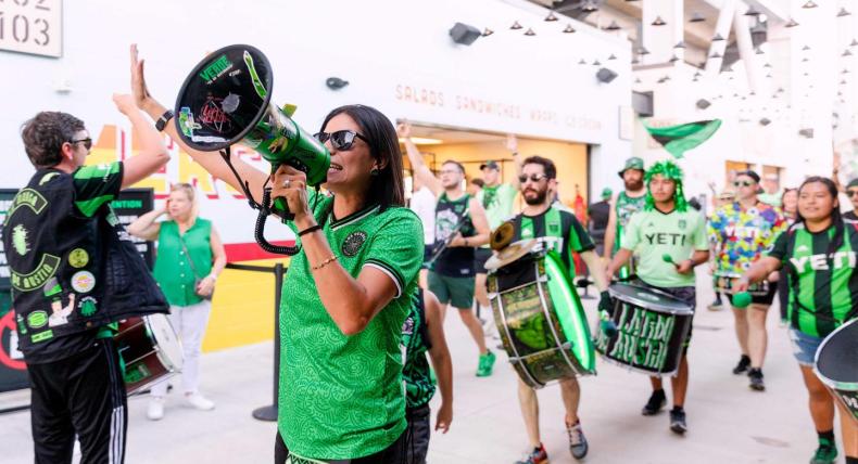 The image shows a group of enthusiastic fans, wearing green and black attire with "YETI" logos, celebrating and marching with drums and a megaphone inside a stadium. The scene is lively, with one person leading chants through the megaphone while others play drums and high-five. The overall atmosphere is energetic and festive, indicating a sports event or rally.