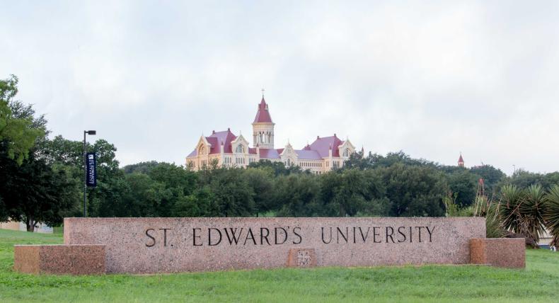 The entrance sign to St. Edward's University, with trees and Main Building in the distance.