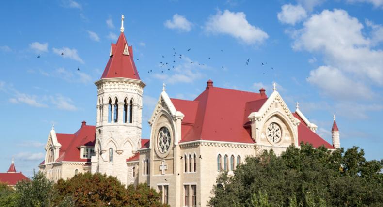 An aerial view of a flock of birds flying over Main Building on a sunny, blue sky day with a few clouds.