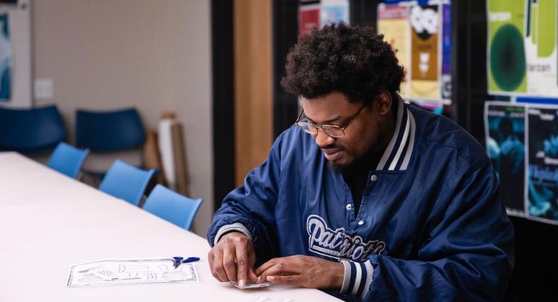 The image shows a man with glasses and a beard, wearing a blue "Patriots" jacket, sitting at a table and concentrating on a craft project involving paper cutouts. He is working alone in a room with blue chairs and colorful posters on the walls, indicating a creative or educational setting.