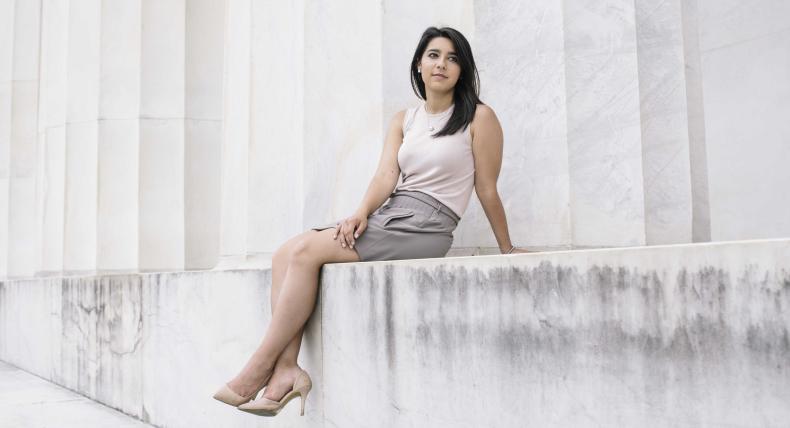 Victoria Ochoa wears a blouse, skirt and heels and sits on a ledge by massive columns.