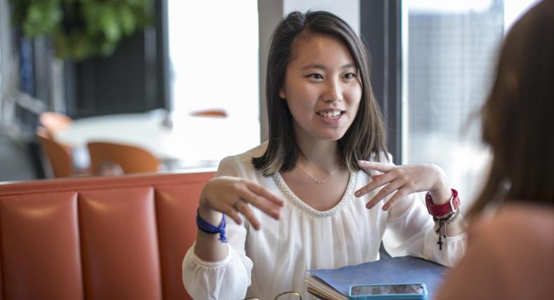 The image shows a young woman with shoulder-length hair, wearing a white blouse with a pearl neckline. She is sitting at a table in a casual setting, engaged in a conversation with another person. She has a blue wristband on her left wrist and a red watch on her right wrist. In front of her are a blue folder and a smartphone. The background includes a large window, some greenery, and blurred elements of a modern café or office space.