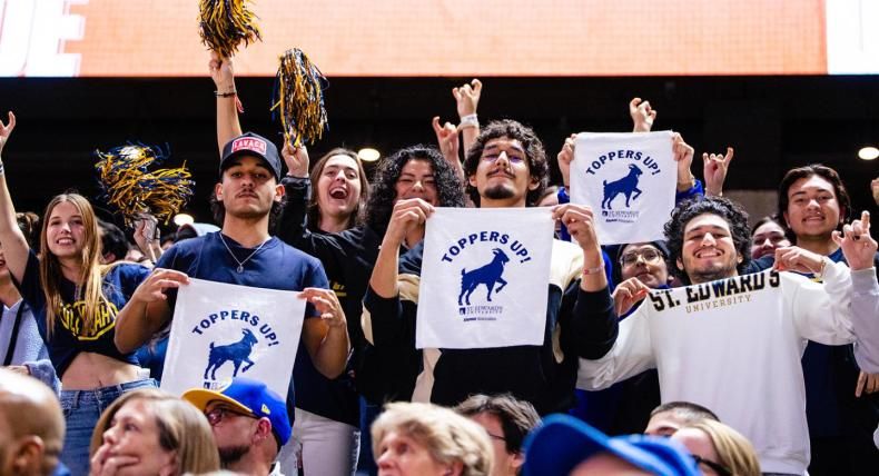 The image depicts a group of individuals at what appears to be a sporting event. They hold up signs or papers with the text "TOPPERS UP" and an image of a horse, suggesting they are fans of a sports team called the Toppers from St. Edwards University. The background includes an orange banner, likely part of the sports venue's decor. Overall, it captures the enthusiasm and community spirit of collegiate athletics.