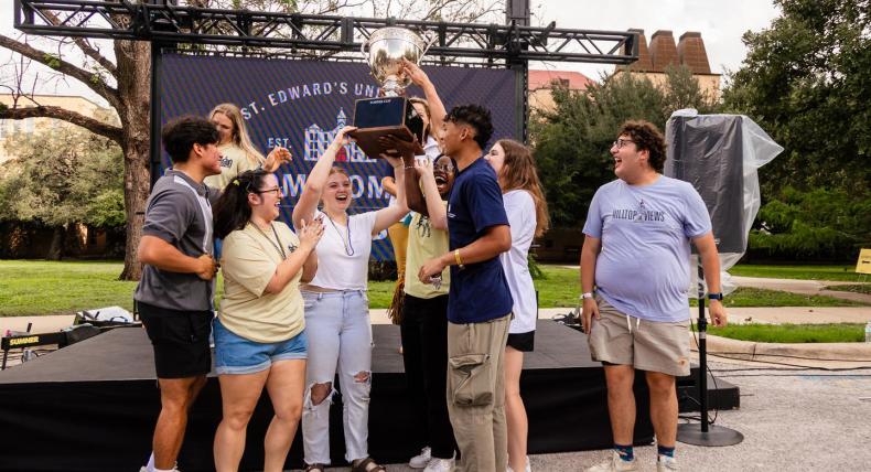 The image shows a group of people standing in front of a banner that reads "St. Edward's University." They are gathered around a trophy, which they are holding up together. Some of them are wearing t-shirts with the word "HILLTOPPERS" on them. It looks like a celebratory moment, possibly after winning a competition or event.