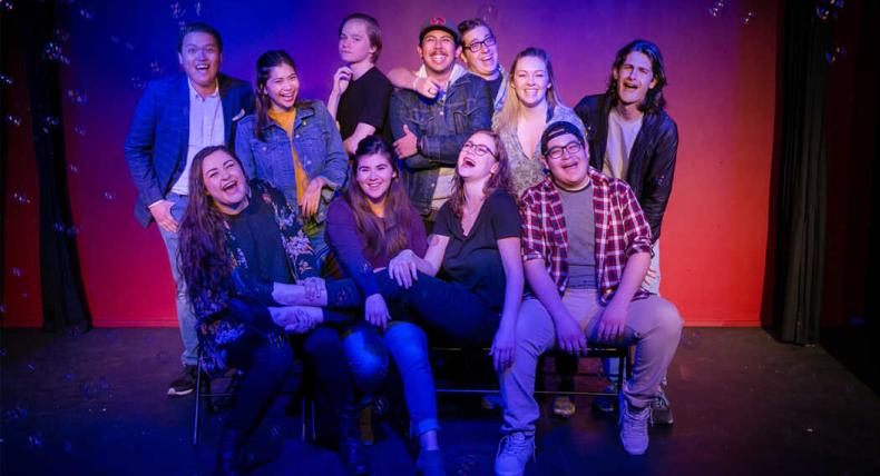 A group of improv students sit and stand and laughter together on a blue and, red and purple background as bubble float around them.