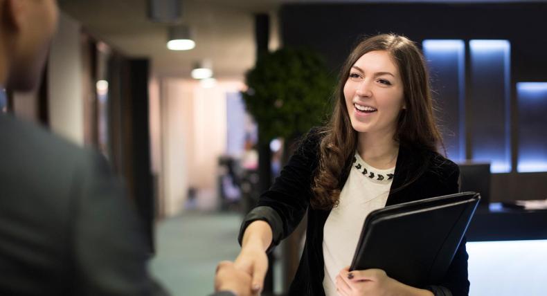 A student wearing a blazer holds a portfolio and shakes hand with their interviewer in an office.