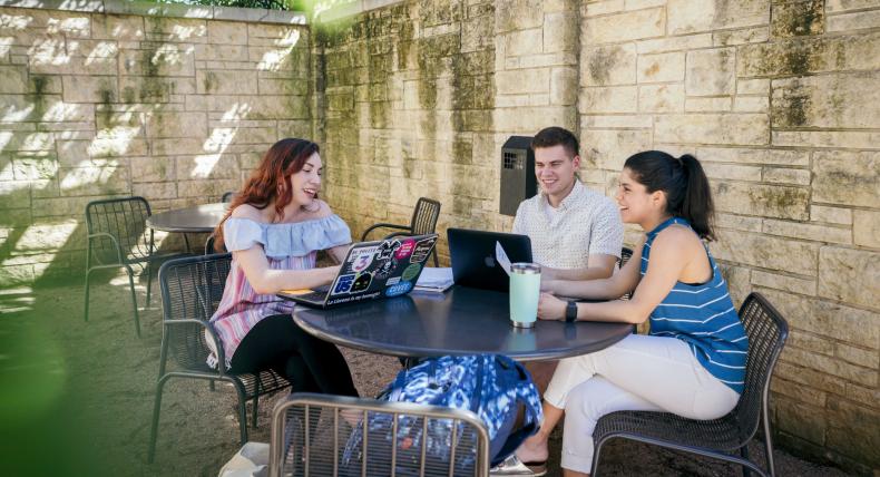 Three students sit at a patio table, hanging out, studying and working on their laptops.