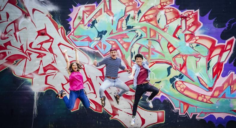 Three students jump in front of a colorful, patterned mural.