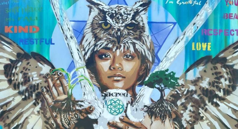 The image is a vibrant mural featuring a young person's face with an owl perched on their head. Their hands hold various elements: a plant, a tree, and a white object with the word "sacred" and a geometric design. The mural incorporates words like "kind," "restful," "love," "respectful," and "beautiful," along with phrases like "you are" and "I'm grateful." The background features a blue and green gradient with geometric shapes, creating a theme of nature, wisdom, and positive affirmations.