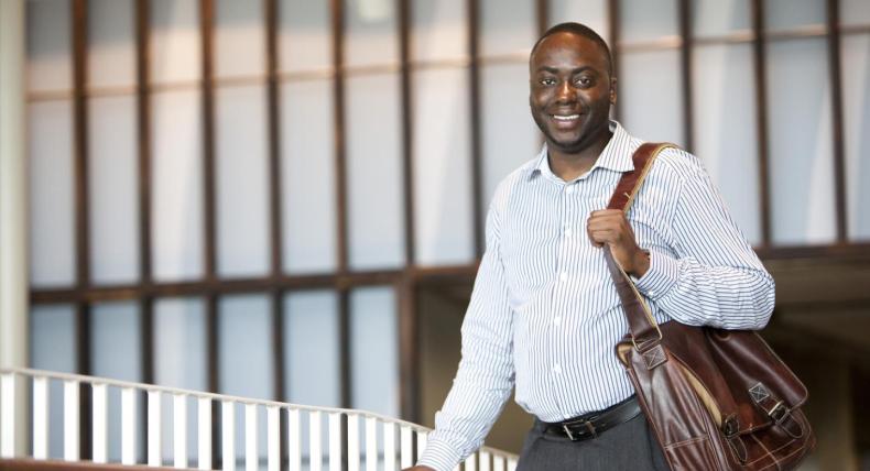 The image shows a smiling man wearing a light blue and white striped button-down shirt and gray trousers. He is carrying a brown leather shoulder bag. The background features a modern interior with large vertical wooden beams and frosted glass panels, giving a professional and contemporary ambiance. The man is positioned near a railing, suggesting he might be in an educational building.