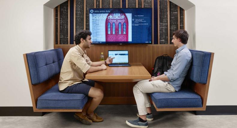 Two students sit in a booth with a table on the table as they look at the screen. A televsion mounted on the wall shows red doors.