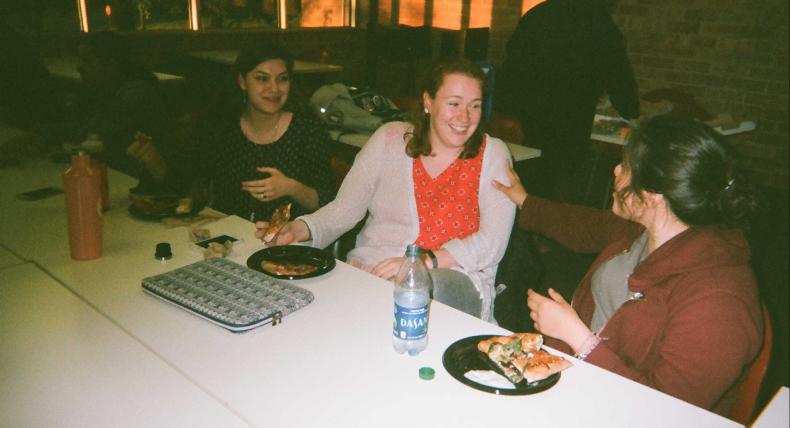 Three students sit at a table with food and talk.