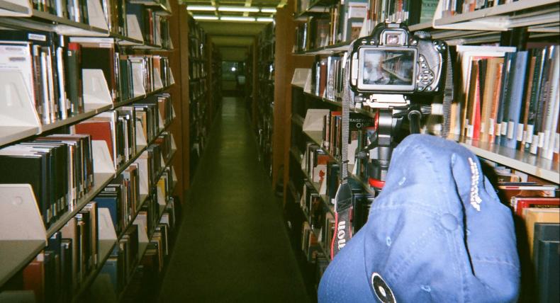 A camera and a hat on a tripod in between book stacks in the Munday Library.