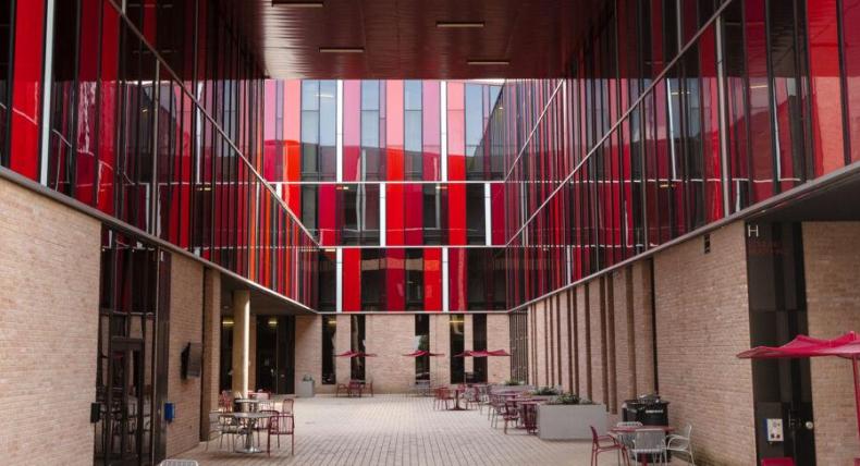 The image depicts a modern, open courtyard surrounded by buildings with striking red and glass facades. The buildings feature vertical red panels in varying shades, creating a dynamic and vibrant appearance. The courtyard is paved with light-colored bricks and is furnished with metal tables and chairs, some with red accents, and red umbrellas. The setting suggests a communal or educational environment, with a clean and contemporary architectural design.