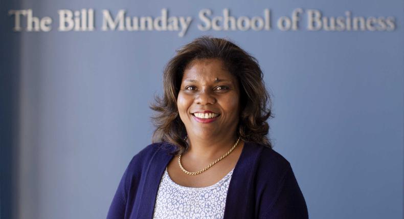 Marianne Ward-Peradoza stands in front of a blue wall that has silver text that reads The Bill Munday School of Business.