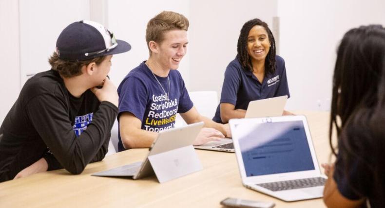 The image shows a group of four young adults sitting around a table, engaged in a lively discussion. They all have laptops in front of them and are smiling or laughing, indicating a positive and collaborative atmosphere. One person is wearing a baseball cap, another is in a branded T-shirt, and a third is wearing a polo shirt with a logo. The setting appears to be a casual meeting or study session in a bright, modern room, suggesting a school or university environment.