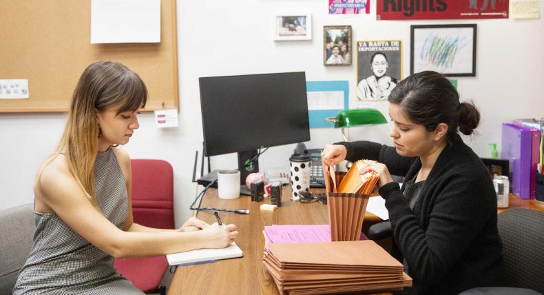 An intern and a staff member work together in an office at a desk, taking notes and filing paperwork.