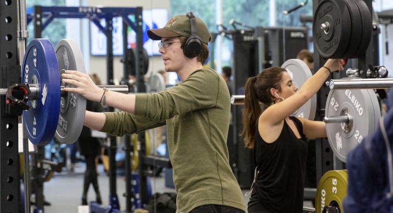 Two students use set up two different barbells on squat racks with weights.