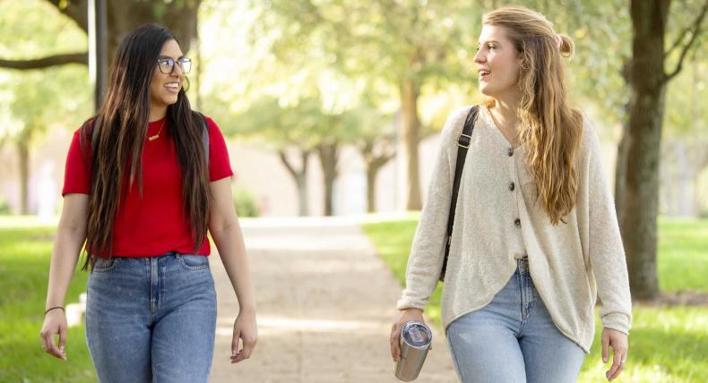 The image shows two young women walking outdoors on a tree-lined path, engaged in conversation. One woman, on the left, is wearing a red T-shirt, jeans, and glasses, while the other, on the right, is dressed in a light-colored cardigan, jeans, and is carrying a reusable tumbler. The scene is bright and sunny, with lush green trees in the background, suggesting a pleasant, casual walk, possibly on a campus or park. The overall mood is friendly and relaxed.