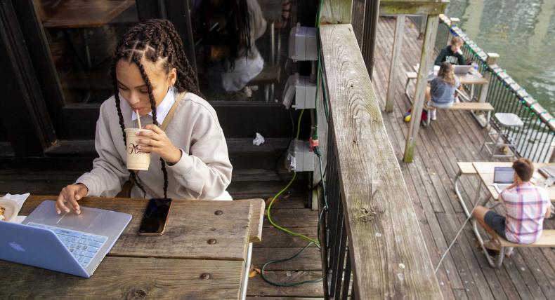 A student sits at a picnic table and drinks coffee while working on their laptop. Other people sitting at tables working on their laptops are seen below.