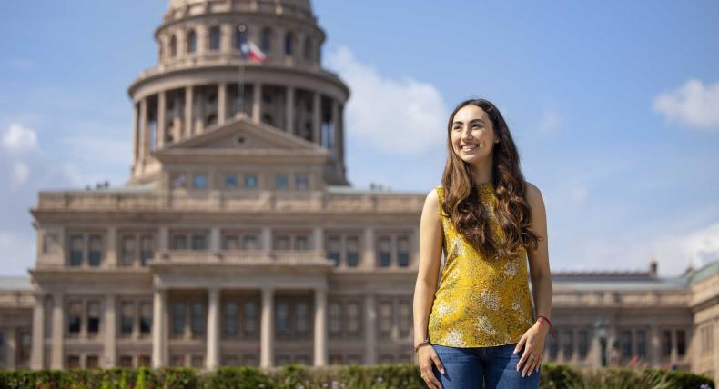 Michelle Flores stands outside of the Texas State Capitol wearing a yellow top and blue jeans on a sunny day.