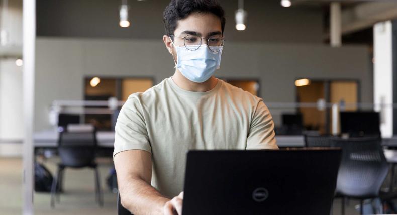 A student wears a face mask and types on a laptop in the Munday Library.