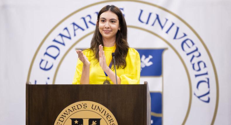 The image shows a woman standing at a podium with the logo of "St. Edward's University" in the background. She is wearing a bright yellow top and is clapping with a smile on her face. The podium has the university's emblem on the front. She appears to be addressing or acknowledging an audience, possibly during a formal event or ceremony at the university.