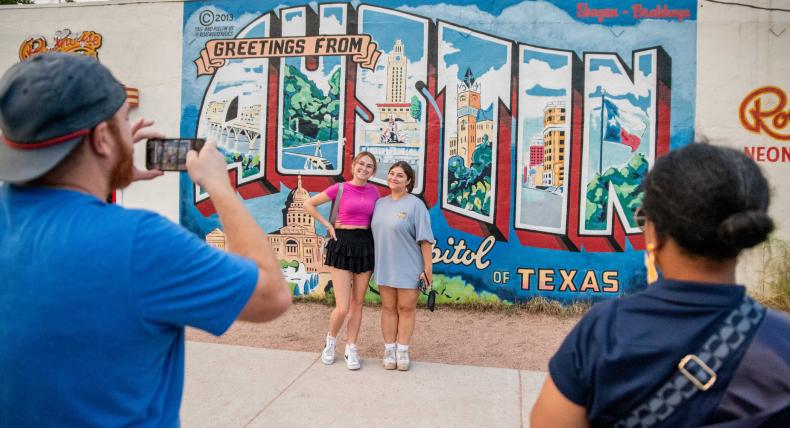 Two students pose in front of the Greetings from Austin mural as two people take their photo in the foreground.