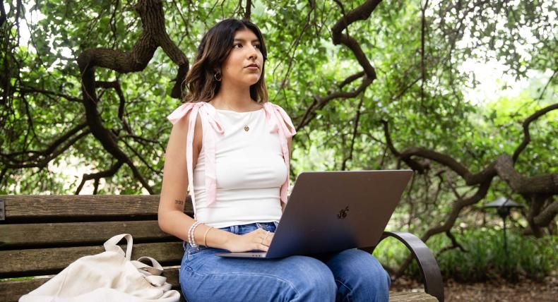 This image features a young woman sitting on a wooden bench in a serene, leafy outdoor setting. She is working on a laptop, with a focused expression, surrounded by twisted branches and green foliage. She wears a white top with pink shoulder ties, blue jeans, and minimal jewelry, including a pendant necklace and bracelets. A light-colored tote bag rests beside her on the bench.