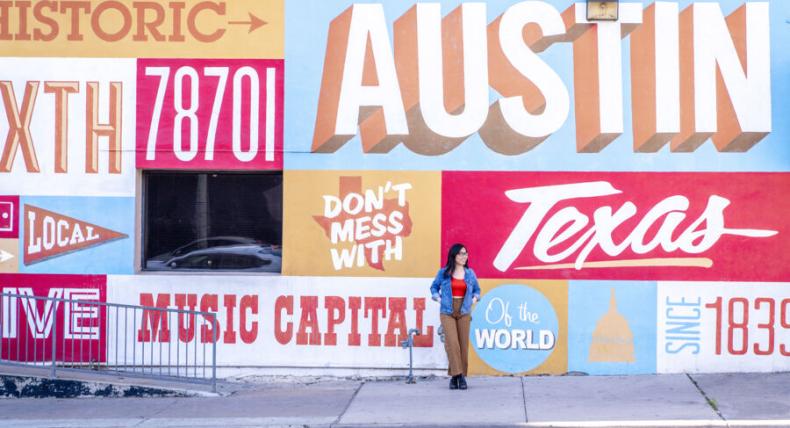 The image shows a large, colorful mural on the side of a building in Austin, Texas. The mural prominently features the words "Austin," "Texas," "Music Capital of the World," and "Don't Mess With Texas," along with other text and graphic elements. A woman is standing in front of the mural, casually posing with one hand on her hip and the other on the wall. She is wearing a denim jacket, red top, and brown pants. The scene captures a vibrant, urban atmosphere.