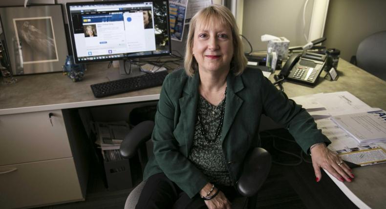 The image shows a woman sitting in an office chair at her desk, smiling at the camera. She is wearing a green blazer and black pants. Her desk is organized, with a computer monitor displaying a professional profile. The workspace includes a phone, papers, and personal items, creating a professional yet personalized environment. The background has various documents and a framed photo, adding to the office's detailed and lived-in feel.