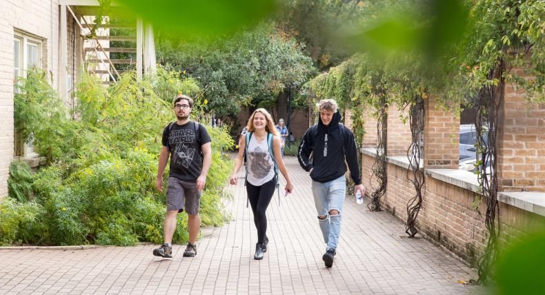 This image shows three young adults walking along a brick-paved path between buildings, surrounded by greenery. The group includes two men and one woman, all casually dressed. The woman in the center is smiling, wearing a backpack, while the man on the left wears a Star Wars t-shirt and the man on the right wears a black hoodie with ripped jeans. The atmosphere is relaxed, with the path framed by plants and vines growing on the walls.