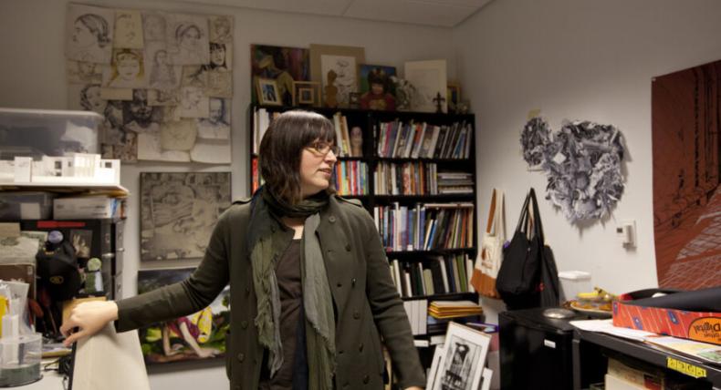 The image shows a woman standing in a cluttered office or studio space. She is wearing a dark coat and a scarf, with glasses and shoulder-length dark hair. The room is filled with bookshelves, art supplies, and various pieces of artwork, including sketches on the walls and framed art on shelves. The woman appears to be in the midst of moving or organizing, as she is looking to the side with her hand resting on a chair. The space has a creative and personal atmosphere.