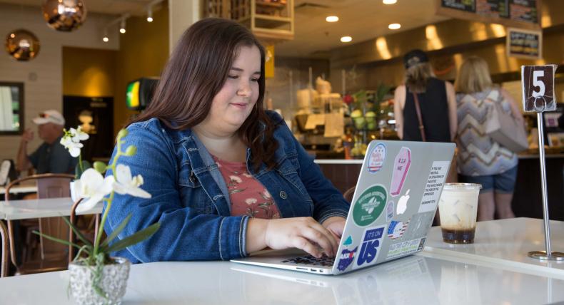 A student types on their laptop and sits at a table in a cafe.