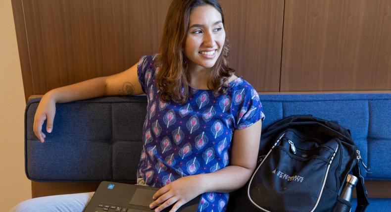 A student sits on a bench with a laptop in their lap and backpack by their side and looks out the window.