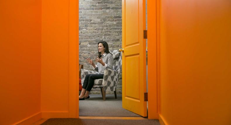 The image shows a woman sitting in an armchair in a room with bright orange walls and an open door. The photo is taken from the hallway, looking into the room through the doorway. The woman is engaged in a conversation, gesturing with her hands, and is wearing a gray sweater and black pants. The room has a brick wall, adding a rustic feel to the modern, brightly colored space. The scene captures a candid moment in what appears to be an informal, cozy setting.