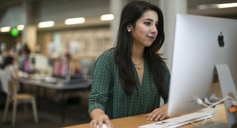 The image shows a woman sitting at a desk, using a large Apple desktop computer. She is holding a computer mouse and looking at the screen with a focused expression. The background appears to be a modern, open-plan office or study space, with other people working at desks in the distance. The woman is wearing a green patterned top, and the environment suggests she might be in a library or co-working space.