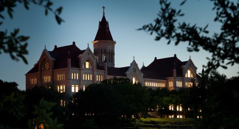 Main Building is framed by foliage in the foreground with windows illuminated at dusk.