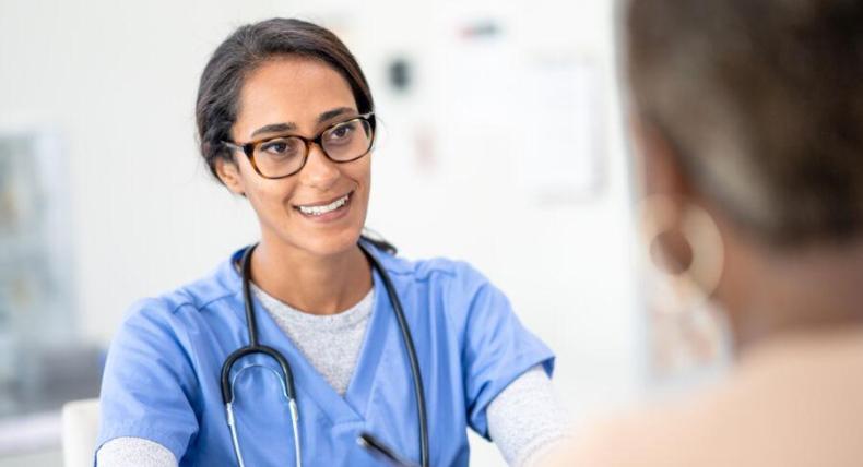 The image shows a healthcare professional, likely a nurse or doctor, engaged in a conversation with a patient. The medical professional is wearing blue scrubs, glasses, and a stethoscope around her neck, and she is smiling warmly, holding a pen and possibly taking notes. The setting appears to be a clinical environment with a bright, clean background. The patient's shoulder is visible in the foreground, with the focus on the healthcare professional.