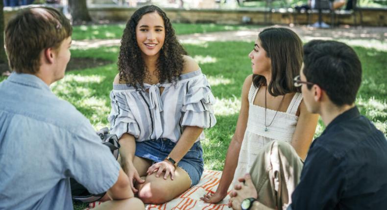 The image shows a group of four young adults sitting on a blanket on a grassy area, likely in a park or on a campus. They are engaged in a casual conversation. The group includes two women and two men. One of the women, with curly hair and wearing an off-the-shoulder top, is smiling as she speaks. The setting is bright and outdoors, with trees and other people in the background, suggesting a relaxed and social environment.