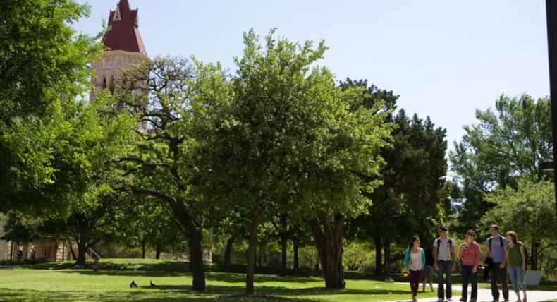 5 students are walking at the St. Edward's Campus with a background that includes trees and the Main Buidling.