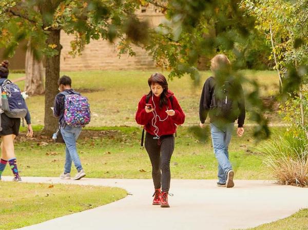 The image shows four students walking along a paved path in a park-like setting with green grass and trees. One student in the foreground is wearing a red jacket and red boots, looking at their phone with earphones on. Two students are walking away from the camera on the left, one wearing colorful socks and carrying a backpack, and the other wearing jeans and a colorful backpack. Another student on the right is also walking away, wearing a black hoodie and jeans.