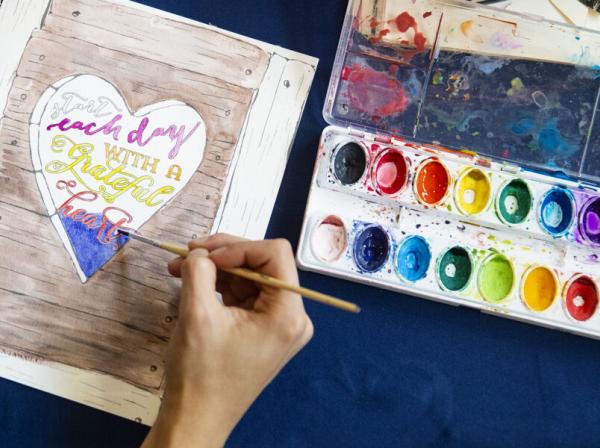 A person paints with watercolors on a page that reads "start each day with a grateful heart" and depicts a heart on wood.