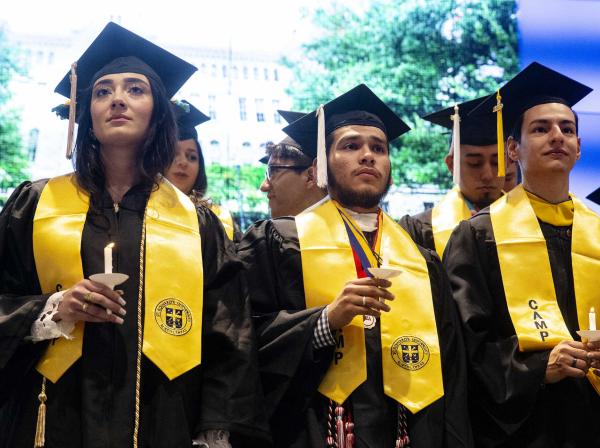 Three CAMP Students hold candles while wearing graduation caps, tassels, robes and stoles. 