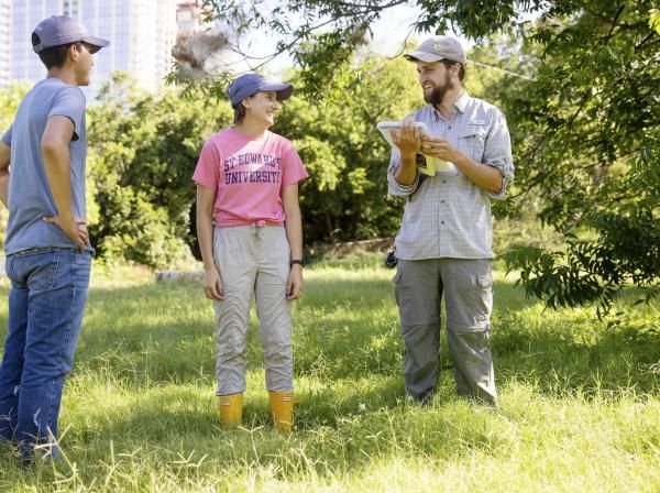 The image shows three people standing in a grassy outdoor area. The person on the left is wearing a blue t-shirt, jeans, and a cap, standing with hands on hips. The person in the middle, wearing a pink "St. Edward's University" t-shirt, beige pants, yellow boots, and a cap, is smiling at the person on the right. The person on the right, wearing a gray shirt, cargo pants, and a cap, is holding a tablet and smiling. The setting is sunny with green trees and a building visible in the background.