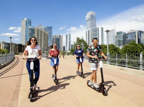 Four students ride scooters acros the Pfluger Pedestrian Bridge with the skyline of Austin in the background.