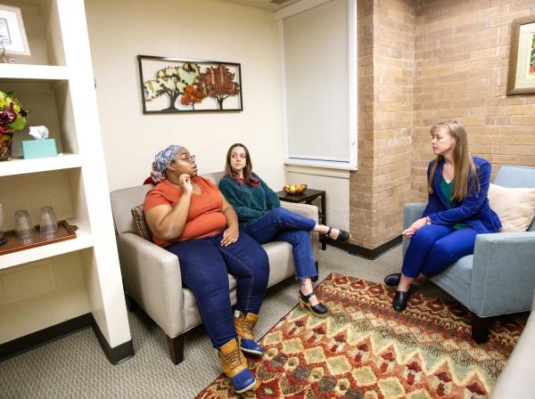 The image depicts three individuals seated in a room. On the left, a person wearing blue jeans and an orange top sits on a beige sofa In the middle, another individual in green pants and a red top occupies a chair with green upholstery. The person on the right, wearing dark pants and a blue top, sits on a light blue armchair. The room features decorative elements like paintings, flowers, and a patterned area rug.