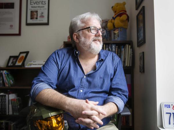 The image shows a man with gray hair and a beard, wearing glasses and a blue button-up shirt, sitting in a room. He is looking to the side with a thoughtful expression. Behind him, there are bookshelves filled with books and various items, including a stuffed Winnie the Pooh bear wearing a cape. On the wall, there are framed pictures and a sign that reads "Events" with a name and photo. The setting appears to be an office or study.